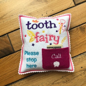 May include: A white square felt pillow with scalloped pink edges, embroidered with the text tooth fairy, a crown, a wand, and stars. It features a dark pink pocket labeled Cali with a tooth icon and the text Please stop here.