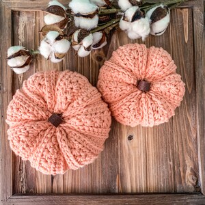 May include: Two crocheted pumpkins in a peach color with brown stems. The pumpkins are sitting on a wooden surface with cotton bolls in the background.