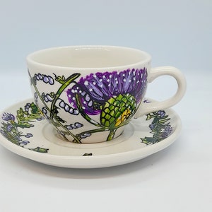 May include: A white teacup and saucer set with a floral design featuring purple and green thistles.