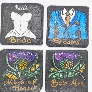 Può includere: Quattro sottobicchieri in ardesia nera con disegni dipinti. Il primo sottobicchiere ha un abito da sposa bianco con una collana di perle e la parola "Bride" in lettere dorate. Il secondo sottobicchiere ha un abito blu con una camicia bianca, una cravatta dorata e la parola "Groom" in lettere dorate. Il terzo sottobicchiere ha un cardo viola e verde con le parole "Maid of Honor" in lettere dorate. Il quarto sottobicchiere ha un cardo viola e verde con le parole "Best Man" in lettere dorate.
