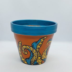 May include: A terracotta flower pot painted with a blue rim and an orange background. The pot is decorated with a yellow and blue octopus design.