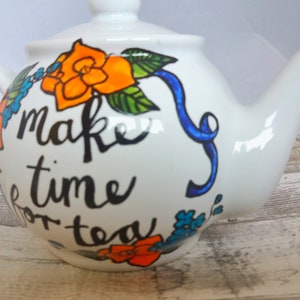 Pu&ograve; includere: Teiera in ceramica bianca con un nastro blu e un motivo floreale. La teiera ha la scritta "Make time for tea".