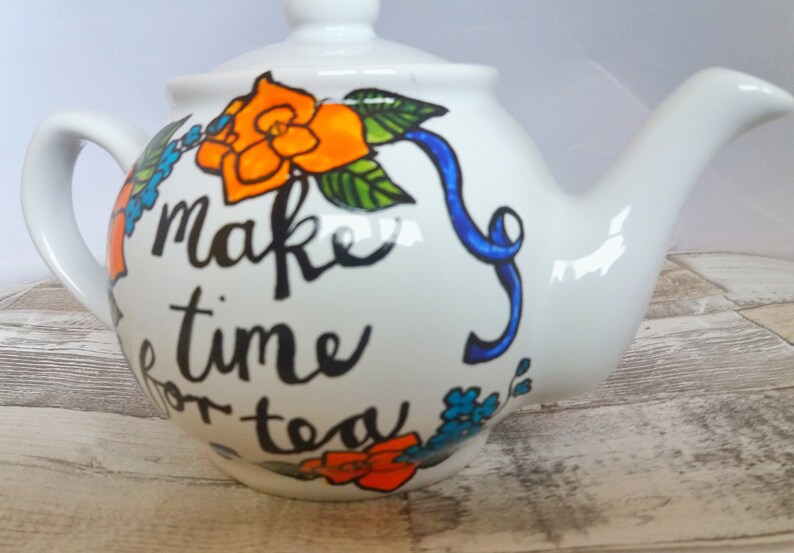 Pu&ograve; includere: Teiera in ceramica bianca con un nastro blu e un motivo floreale. La teiera ha la scritta "Make time for tea".