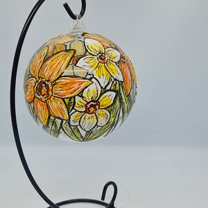 Op de afbeelding: Een glazen ornament met een zwarte metalen standaard, met handgeschilderde gele en witte narcissen.