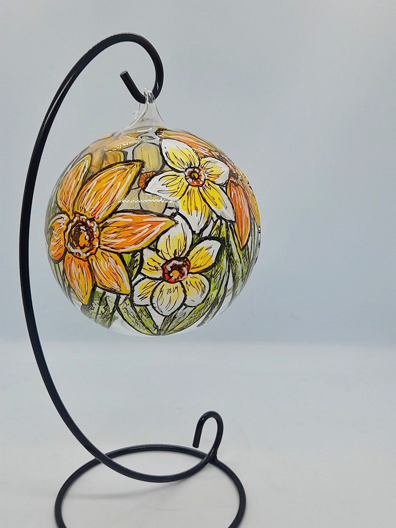 Op de afbeelding: Een glazen ornament met een zwarte metalen standaard, met handgeschilderde gele en witte narcissen.