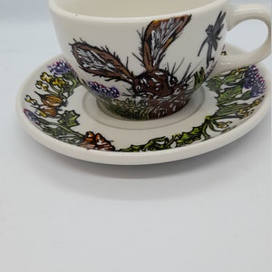 May include: A white ceramic teacup and saucer with a hand-painted design of a rabbit, flowers, and a dragonfly. The rabbit is brown and white, and the flowers are purple, yellow, and orange.