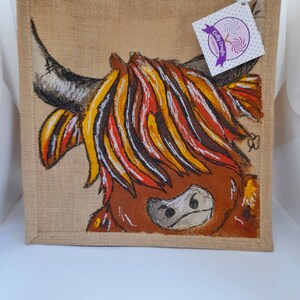 May include: A jute tote bag featuring a hand-painted Highland cow. The cow has a brown face, black horns, and a colorful mane of yellow, orange, and white. A tag with the text "Lolly Crafts Creations" is attached.