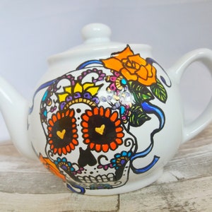 Pu&ograve; includere: Teiera in ceramica bianca con un designo di teschio di zucchero dipinto a mano. Il teschio presenta dettagli in arancione, nero, giallo e viola. La teiera ha un nastro blu e un fiore giallo sul lato.