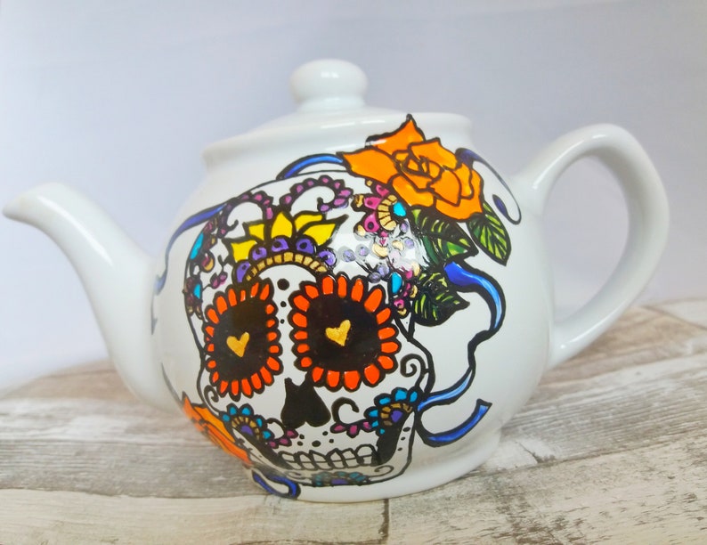 Pu&ograve; includere: Teiera in ceramica bianca con un designo di teschio di zucchero dipinto a mano. Il teschio presenta dettagli in arancione, nero, giallo e viola. La teiera ha un nastro blu e un fiore giallo sul lato.