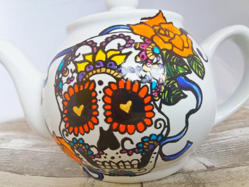 Pu&ograve; includere: Teiera in ceramica bianca con un design di teschio di zucchero dipinto a mano. Il teschio &egrave; decorato con fiori colorati, cuori e volute. La teiera ha un nastro blu e un fiore giallo sulla parte superiore.
