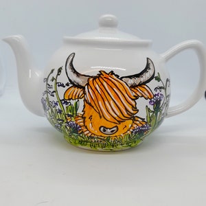 Peut inclure: Une théière en céramique blanche avec un motif peint à la main d'une vache des Highlands dans un champ de fleurs sauvages. La vache est orange et blanche avec un nez noir et des sabots noirs. Les fleurs sauvages sont violettes, vertes et jaunes.