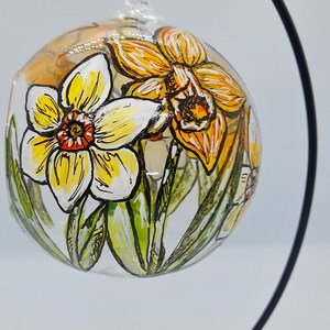 Op de afbeelding: Een glazen ornament met een handgeschilderd ontwerp van gele en oranje narcissen. Het ornament hangt aan een zwarte metalen standaard.