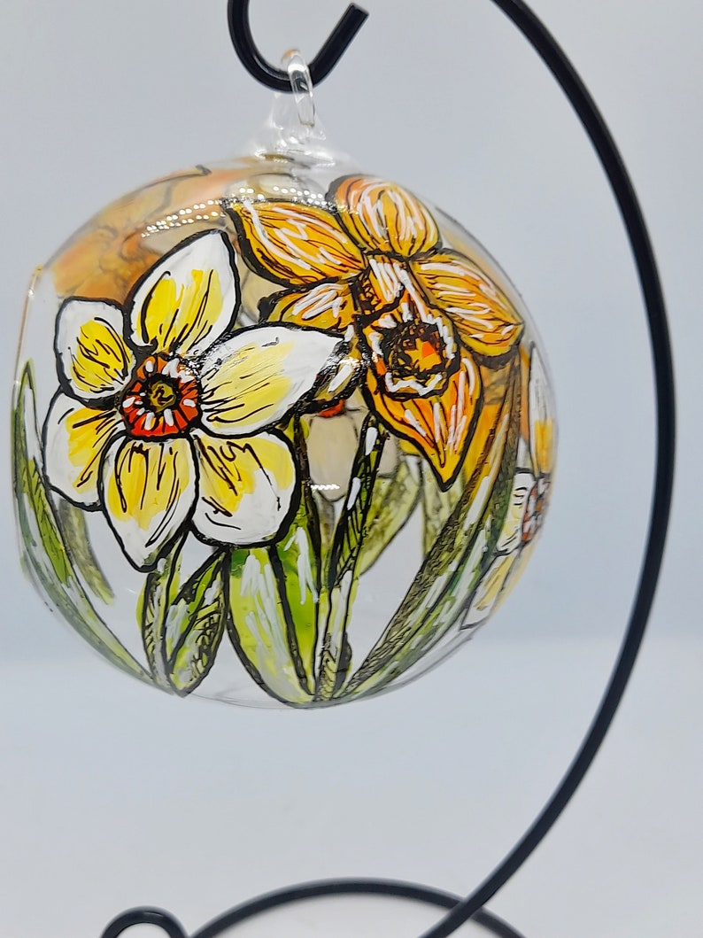 Op de afbeelding: Een glazen ornament met een handgeschilderd ontwerp van gele en oranje narcissen. Het ornament hangt aan een zwarte metalen standaard.