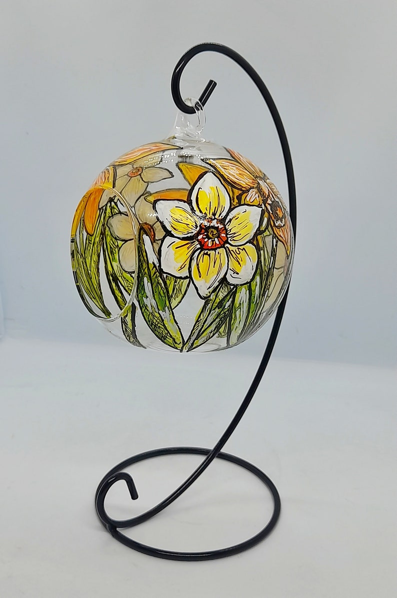 Op de afbeelding: Een glazen ornament met een zwarte metalen standaard, met handgeschilderde gele narcissen met groene bladeren en oranje centra.