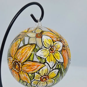 Op de afbeelding: Een glazen ornament met een handgeschilderd ontwerp van gele en oranje narcissen. Het ornament hangt aan een zwarte metalen standaard.