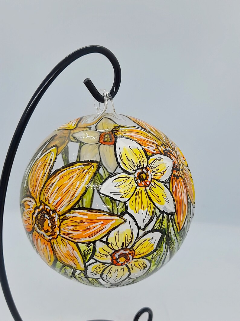 Op de afbeelding: Een glazen ornament met een handgeschilderd ontwerp van gele en oranje narcissen. Het ornament hangt aan een zwarte metalen standaard.