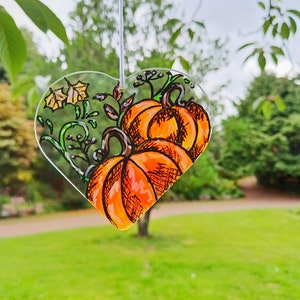 Puede incluir: Un adorno de vidrio en forma de corazón con un diseño pintado a mano de dos calabazas naranjas y enredaderas verdes. Las calabazas tienen contornos negros y están pintadas con colores naranja, amarillo y verde.