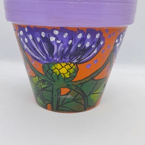 May include: A terracotta flower pot with a purple and white painted thistle design. The pot has a light purple rim and a bright orange background.