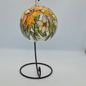 Op de afbeelding: Een heldere glazen bal ornament met een zwarte metalen standaard. De ornament is handgeschilderd met gele, oranje en witte bloemen.