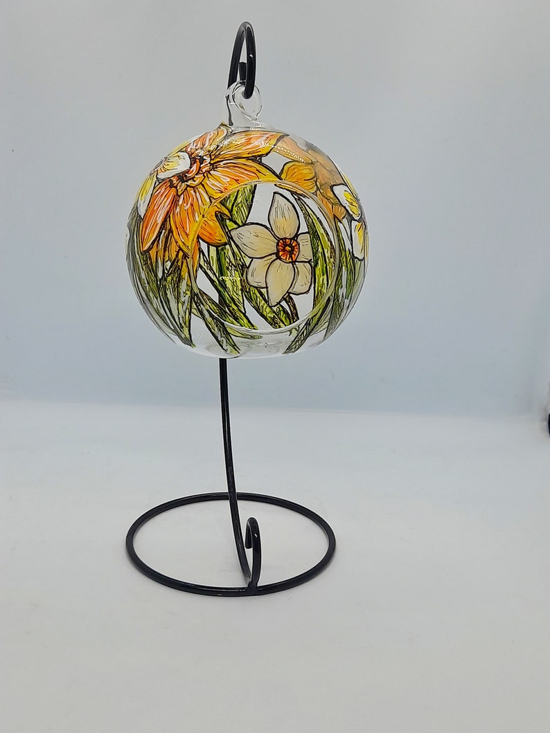 Op de afbeelding: Een heldere glazen bal ornament met een zwarte metalen standaard. De ornament is handgeschilderd met gele, oranje en witte bloemen.