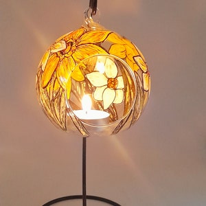 Op de afbeelding: Een glazen kaarshouder met een bloemmotief met gele en witte narcissen. De houder staat op een zwarte metalen standaard met een ronde basis.