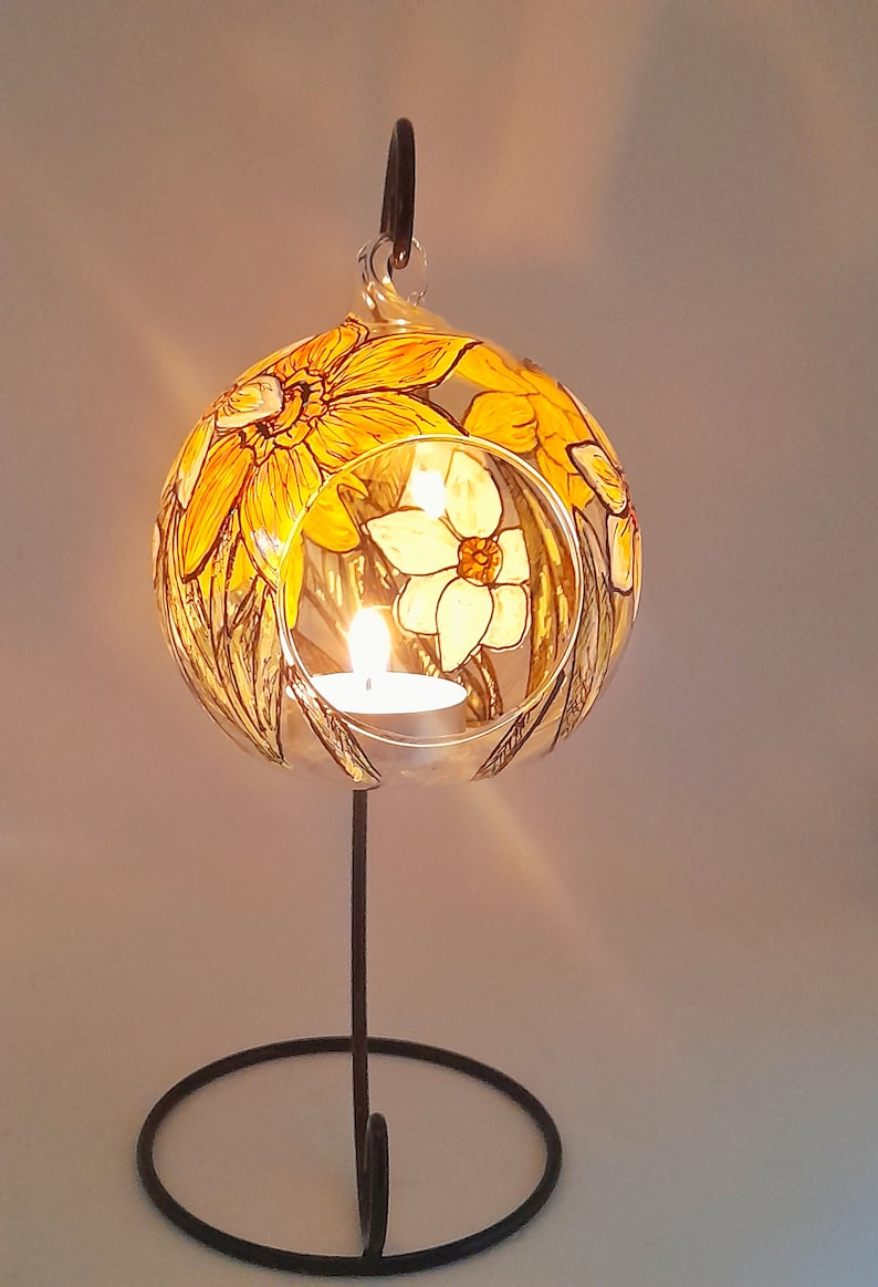 Op de afbeelding: Een glazen kaarshouder met een bloemmotief met gele en witte narcissen. De houder staat op een zwarte metalen standaard met een ronde basis.