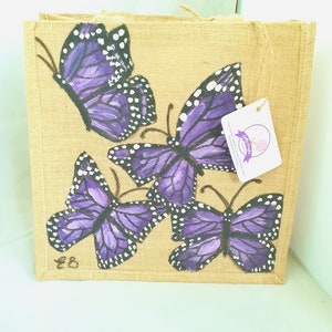 May include: A reusable burlap tote bag with four purple butterflies painted on it. The butterflies have black outlines and white spots. The bag has a brown handle and the text "$6" is written on the bottom left corner.