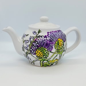 May include: White ceramic teapot with a floral design featuring purple and green thistles. The teapot has a lid and a handle.