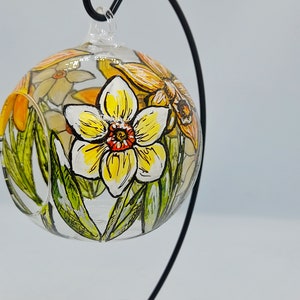 Op de afbeelding: Een glazen bolvormige ornament met een handgeschilderd ontwerp van gele narcissen en groene bladeren. De ornament hangt aan een zwarte metalen standaard.