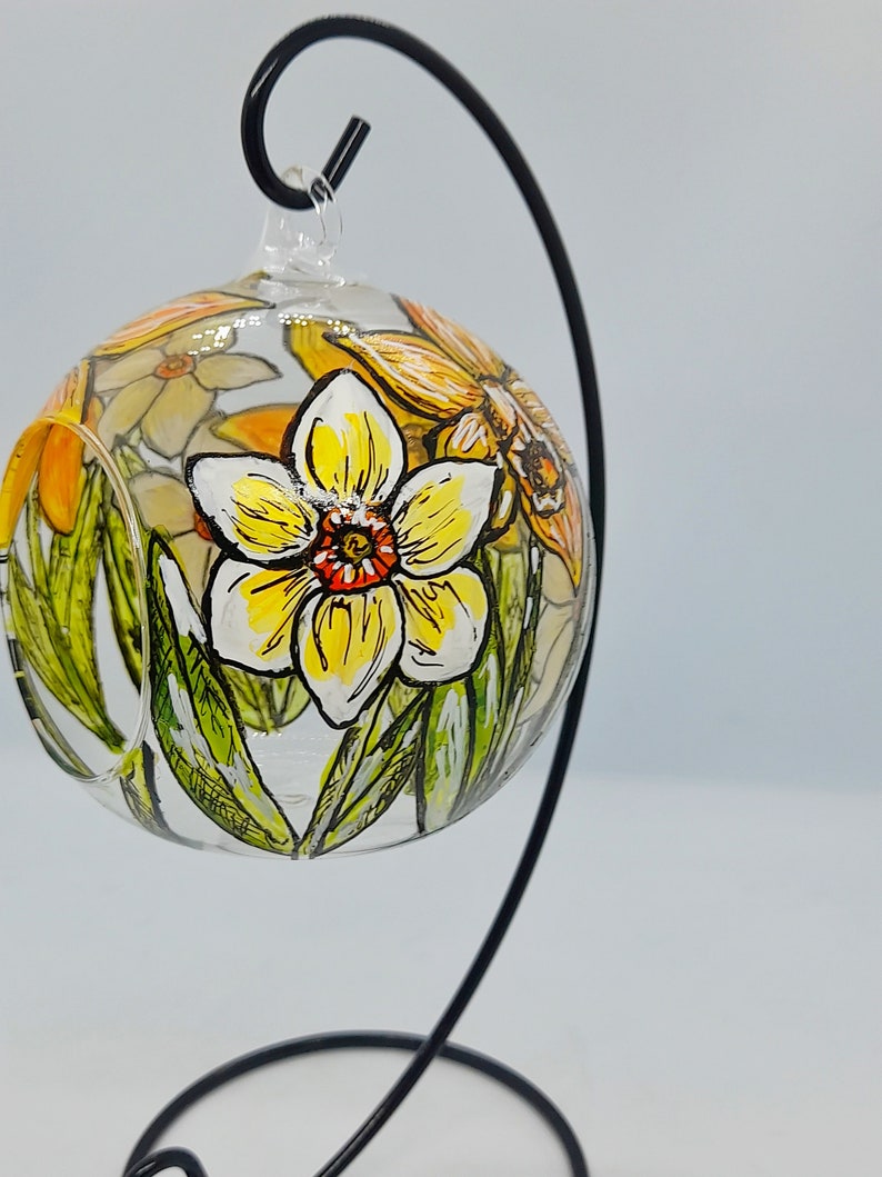 Op de afbeelding: Een glazen bolvormige ornament met een handgeschilderd ontwerp van gele narcissen en groene bladeren. De ornament hangt aan een zwarte metalen standaard.