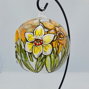 Op de afbeelding: Een glazen bol ornament met een handgeschilderd ontwerp van gele en oranje narcissen. De bol hangt aan een zwarte metalen standaard.