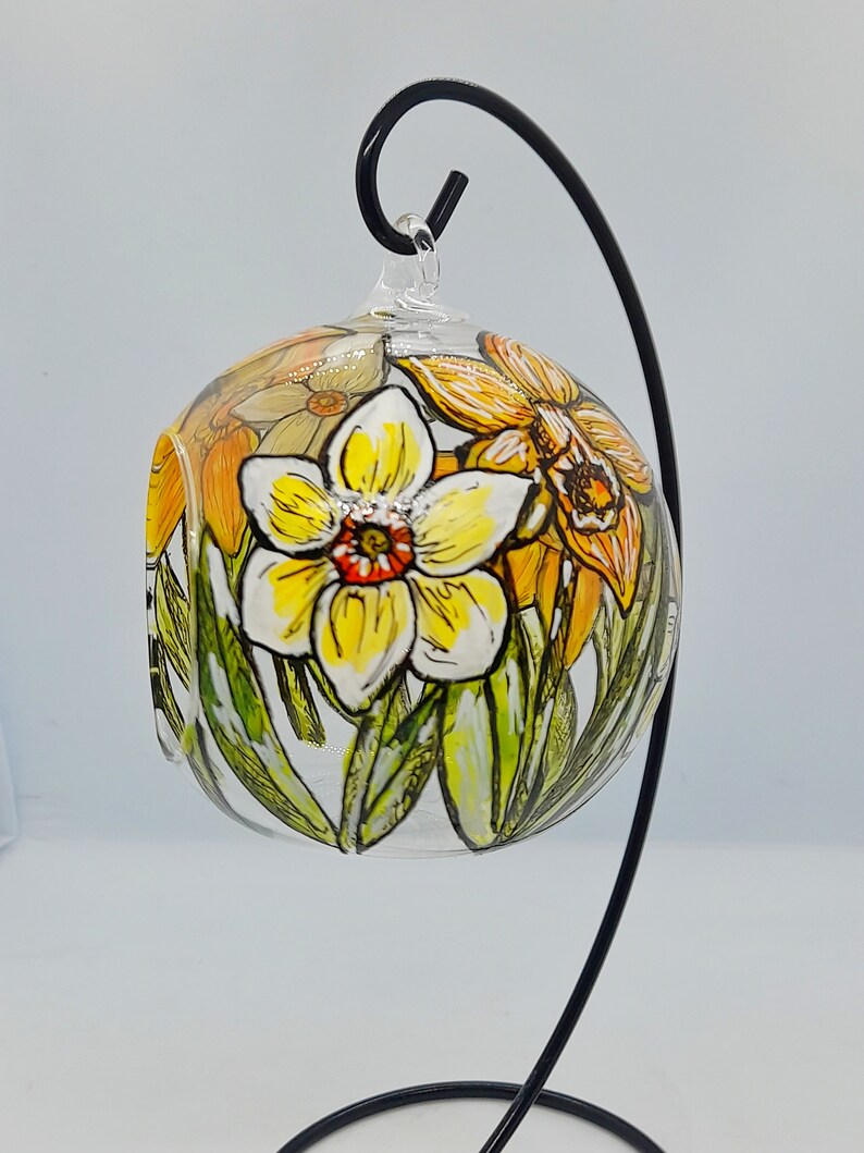 Op de afbeelding: Een glazen bol ornament met een handgeschilderd ontwerp van gele en oranje narcissen. De bol hangt aan een zwarte metalen standaard.