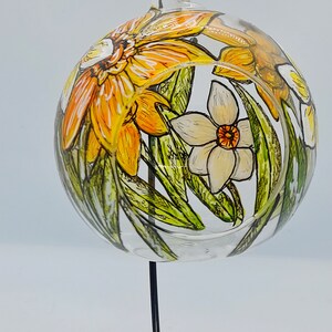 Op de afbeelding: Een glazen bolvormige ornament met een handgeschilderd ontwerp van gele en witte narcissen met groene bladeren. De ornament hangt aan een zwarte metalen standaard.
