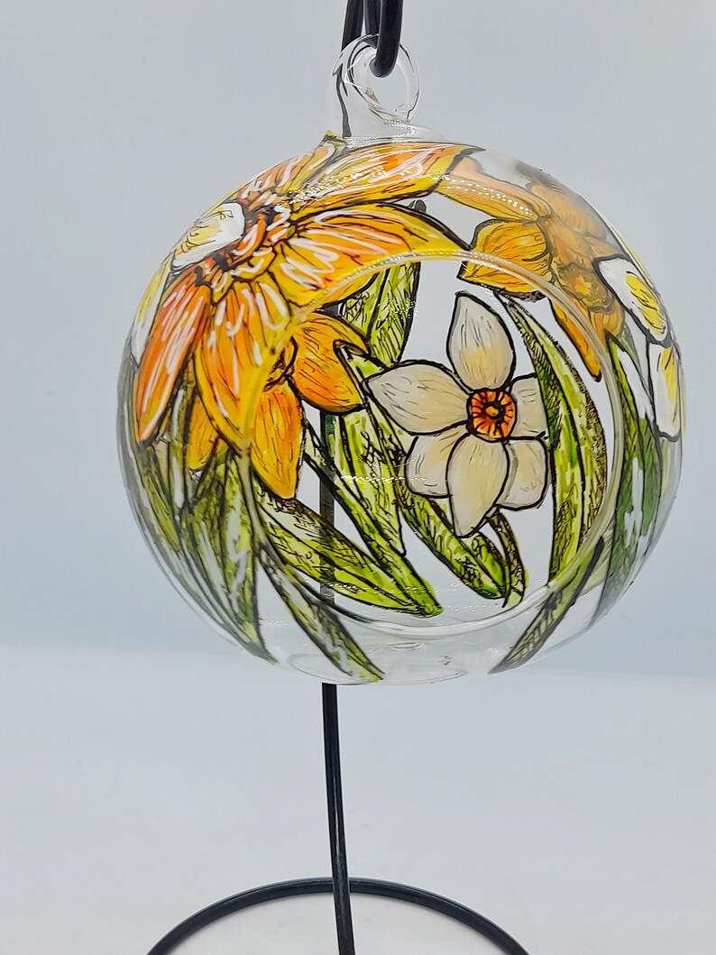 Op de afbeelding: Een glazen bolvormige ornament met een handgeschilderd ontwerp van gele en witte narcissen met groene bladeren. De ornament hangt aan een zwarte metalen standaard.