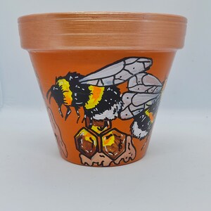 May include: A terracotta flower pot decorated with a painted bumblebee and honeycomb design. The bee is black and yellow with white wings. The honeycomb is brown and yellow.