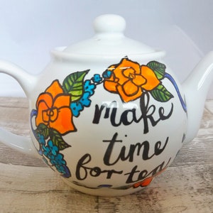 Pu&ograve; includere: Teiera bianca con un design floreale e il testo "make time for tea". I fiori sono arancioni, blu e verdi.