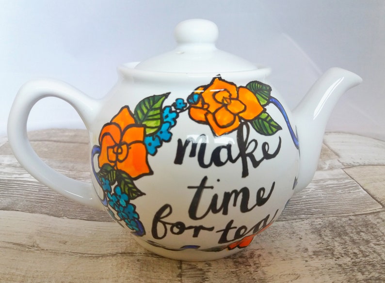 Pu&ograve; includere: Teiera bianca con un design floreale e il testo "make time for tea". I fiori sono arancioni, blu e verdi.