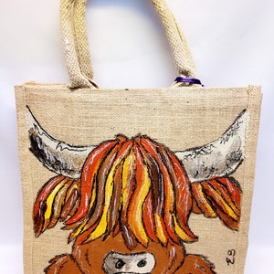 May include: A jute tote bag with a hand-painted Highland cow design. The cow features a shaggy, brown body, a white face, and large horns. The bag has rope handles and a natural, tan color.