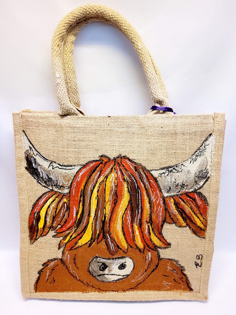 May include: A jute tote bag with a hand-painted Highland cow design. The cow features a shaggy, brown body, a white face, and large horns. The bag has rope handles and a natural, tan color.