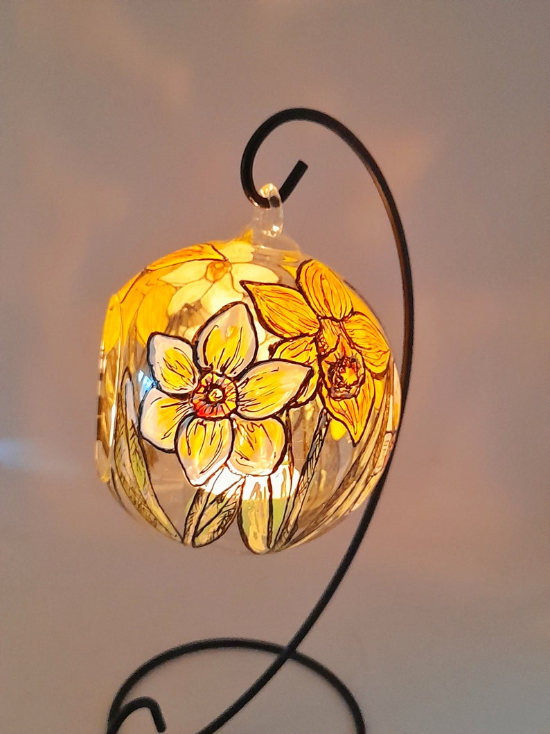 Op de afbeelding: Een glazen bol met een zwarte metalen standaard, handgeschilderd met gele narcissen. De bol wordt van binnenuit verlicht, waardoor een warme gloed ontstaat.