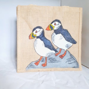 Puede incluir: Una bolsa de lona marrón con dos frailecillos pintados a mano sobre una roca gris. Los frailecillos son blancos con marcas negras y azules y picos naranjas.