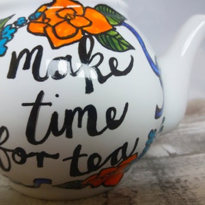 Pu&ograve; includere: Teiera bianca con una scritta dipinta a mano in nero che dice "make time for tea". La teiera &egrave; decorata con fiori arancioni e blu.