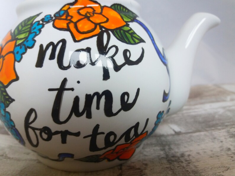 Pu&ograve; includere: Teiera bianca con una scritta dipinta a mano in nero che dice "make time for tea". La teiera &egrave; decorata con fiori arancioni e blu.