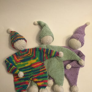 Puede incluir: Tres muñecas de punto con pompones blancos en sus gorros. Las muñecas llevan trajes de rayas de colores, verde y morado.