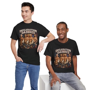 Puede incluir: Una camiseta negra con un estampado gráfico de cuatro figuras históricas musculosas y el texto DECLARATION DADDIES encima. El diseño incluye un fondo con patrones de la bandera estadounidense y fuegos artificiales, con los personajes portando mosquetes.