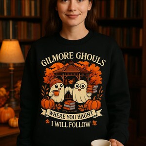 Peut inclure: Un sweat-shirt noir à col rond avec un motif représentant deux fantômes dans un kiosque, entourés de citrouilles et de livres. Le texte indique GILMORE GHOULS WHERE YOU HAUNT I WILL FOLLOW. Le design inclut des feuilles d'automne orange.