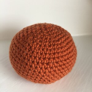 May include: A round, orange crocheted ball. The ball is made of a textured yarn and has a dense, solid appearance. The color is a warm, earthy orange, and the stitches are clearly visible, giving it a handmade look.