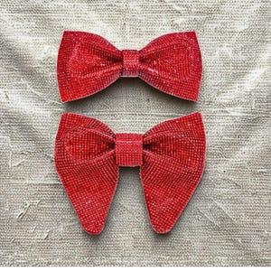 May include: Two red, glitter-covered bow accessories. The bows are identical in shape, with a classic bow tie design. The glitter catches the light, creating a sparkling effect. The bows are set against a neutral, textured background.
