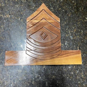 Puede incluir: Una placa de madera con un diseño tallado de una insignia de rango militar. La insignia es una forma de chevron con cinco rayas y un diamante en el centro. La placa está hecha de madera marrón claro y tiene un acabado liso.