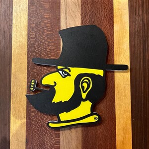 May include: A black and yellow graphic of a man wearing a hat and smoking a pipe. The man has a black beard and mustache. The graphic is on a wooden cutting board.